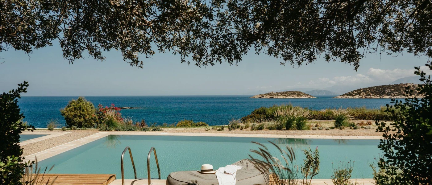 Ocean-facing rectangular swimming pool framed by trees with a grey daybed to one side with a straw hat and towel on