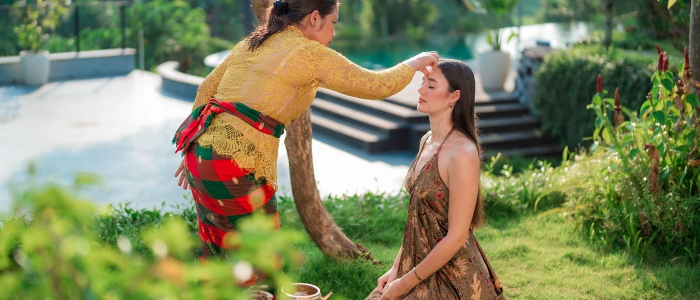 Woman in Balinese dress touching the forehead of a kneeling woman in a tropical garden