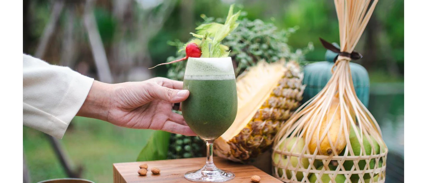 Glass of green juice with a cherry rested on top, next to a basket of fruit and a large pineapple