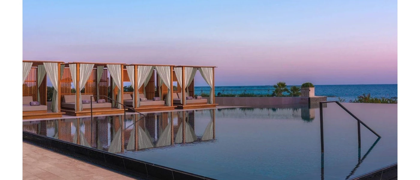 Swimming pool's edge surrounded by four-poster wooden daybeds with white curtains, overlooking the ocean as the sun goes down