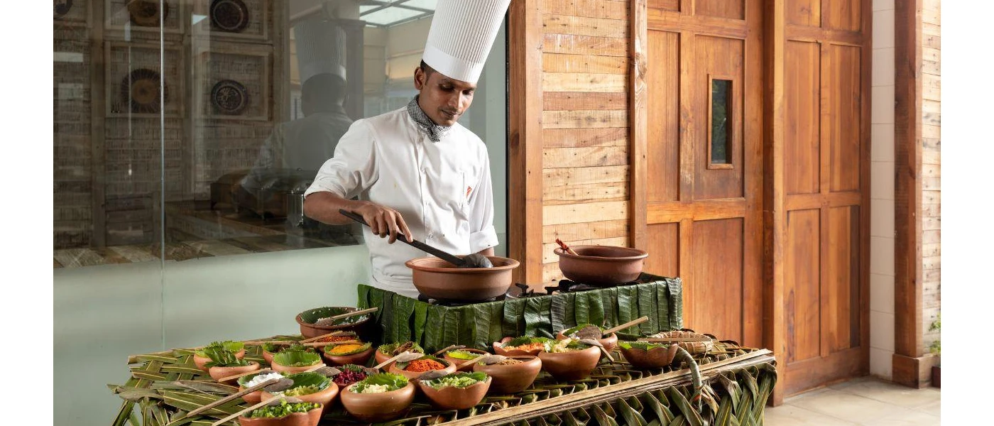Chef in whites prepares food on an outdoor cooking station covered in bamboo leaves and wooden bowls