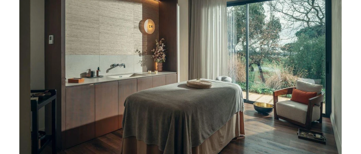 Stylish spa treatment room with wooden flooring and wood panelling, a floor-to-ceiling window, a bed with grey towels on it and a vanity unit with a vase of pink flowers on top