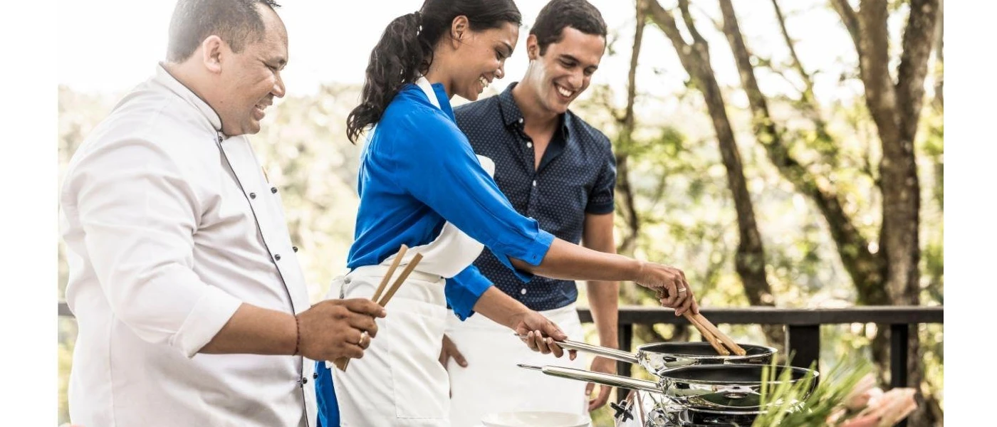Man, woman and chef in whites smiling together as they cook with pans over a hob in the open air