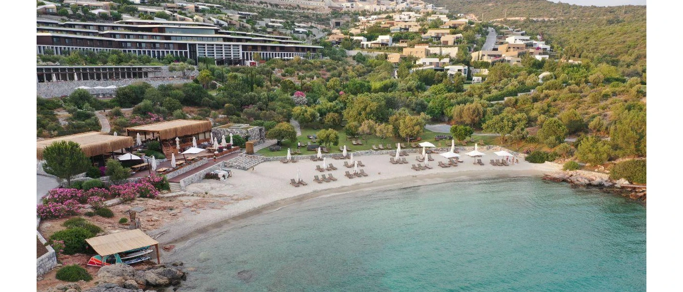 Milky beach furnished with loungers and parasols, surrounded by greenery and overlooked by white buildings