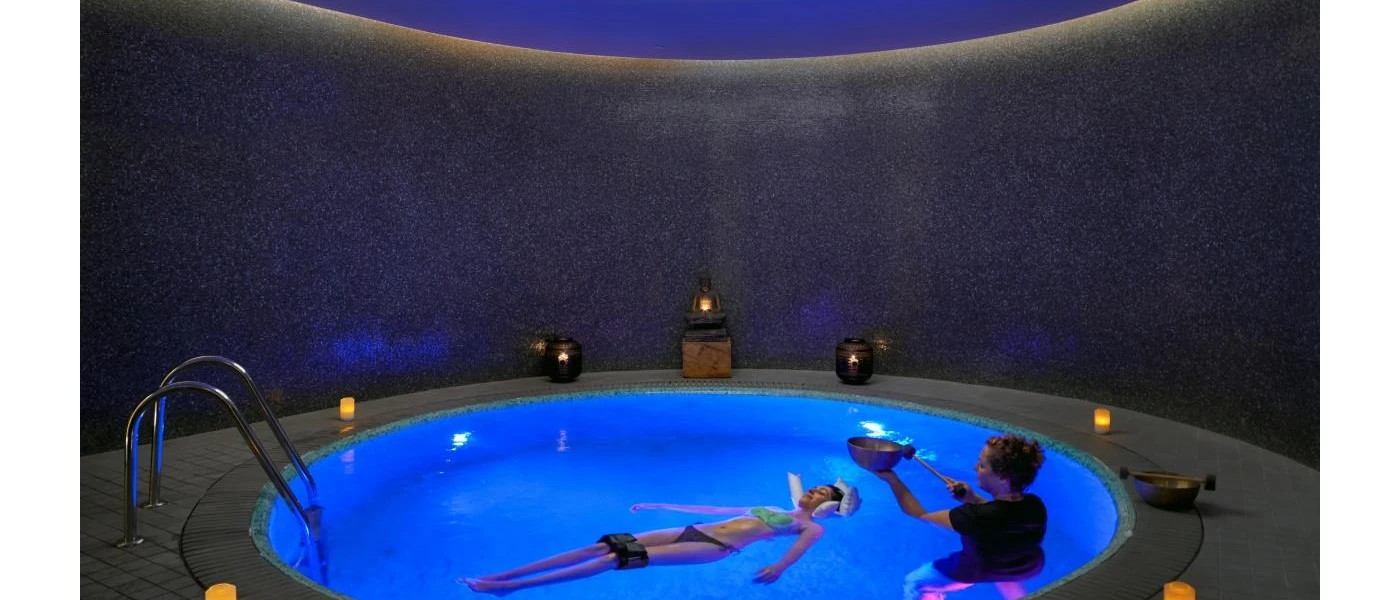 Woman relaxing on her back in a candelit pool as a therapist practices sound healing beside her