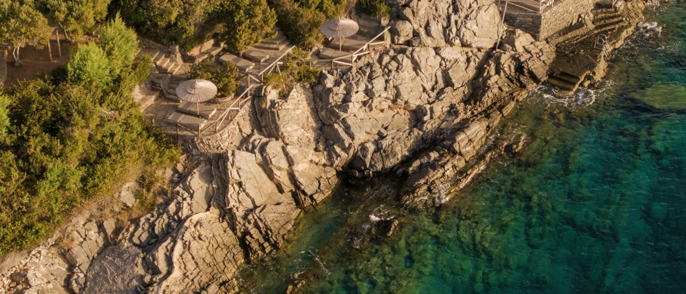Rugged rockface with stone platforms with loungers and umbrellas, overlooking turquoise waves and fringed by greenery