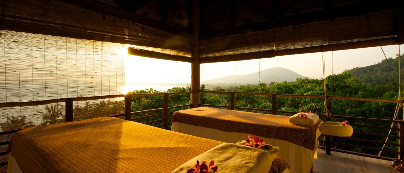 Thatched-roof open-sided treatment cabana with two massage beds covered in yellow fabric and pillows with pink flowers on top and a sea view