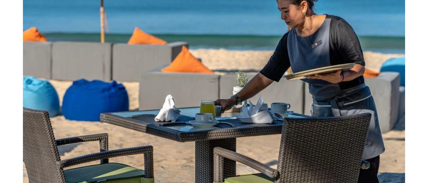 Staff member sets a table on the beachfront, with blue bean bags and orange cushions in the background