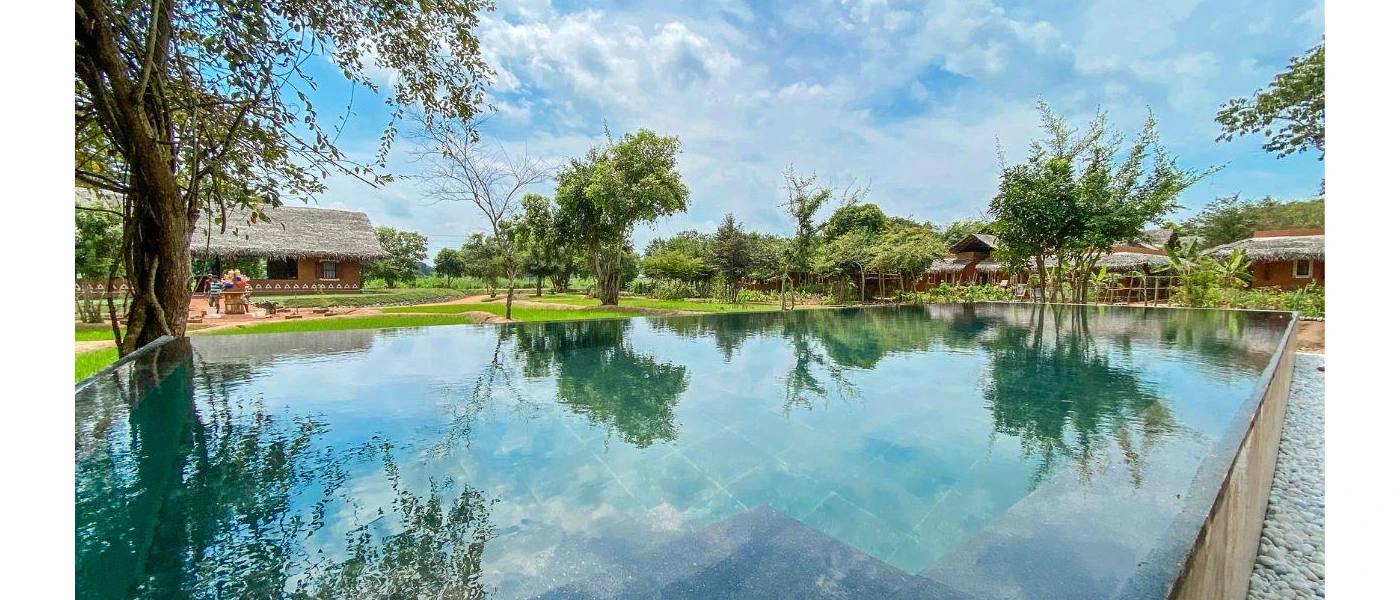 Rectangular swimming pool surrounded by lawns, trees and thatched-roof buildings under a blue sky