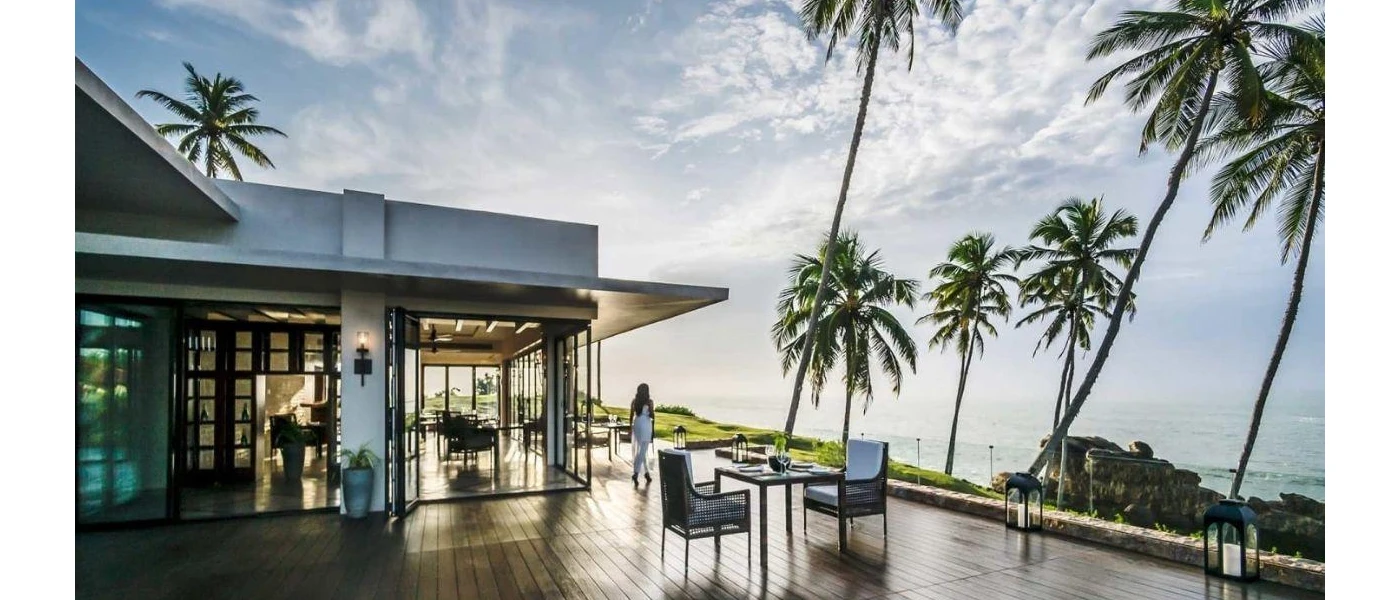 Woman in white walking on a wooden terrace overlooking the ocean, with table and chairs set up for dining