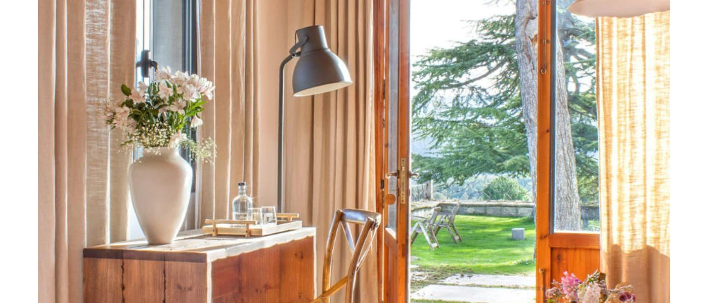 Neutral room with cream curtains, a wooden table with a white vase and flowers, an open balcony door and a view of the gardens