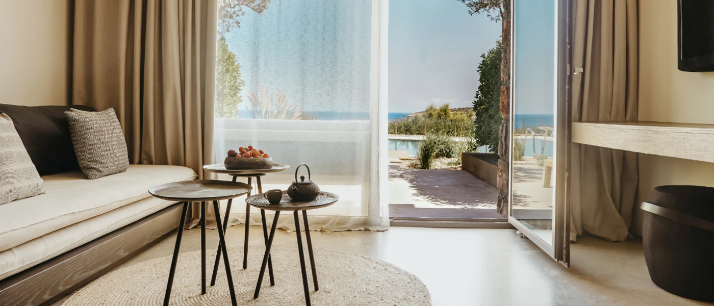 Hotel bedroom living area with a neutral sofa and cushions, round rattan rug, dark circular coffee tables and a wall-hung TV, plus a balcony with sea views