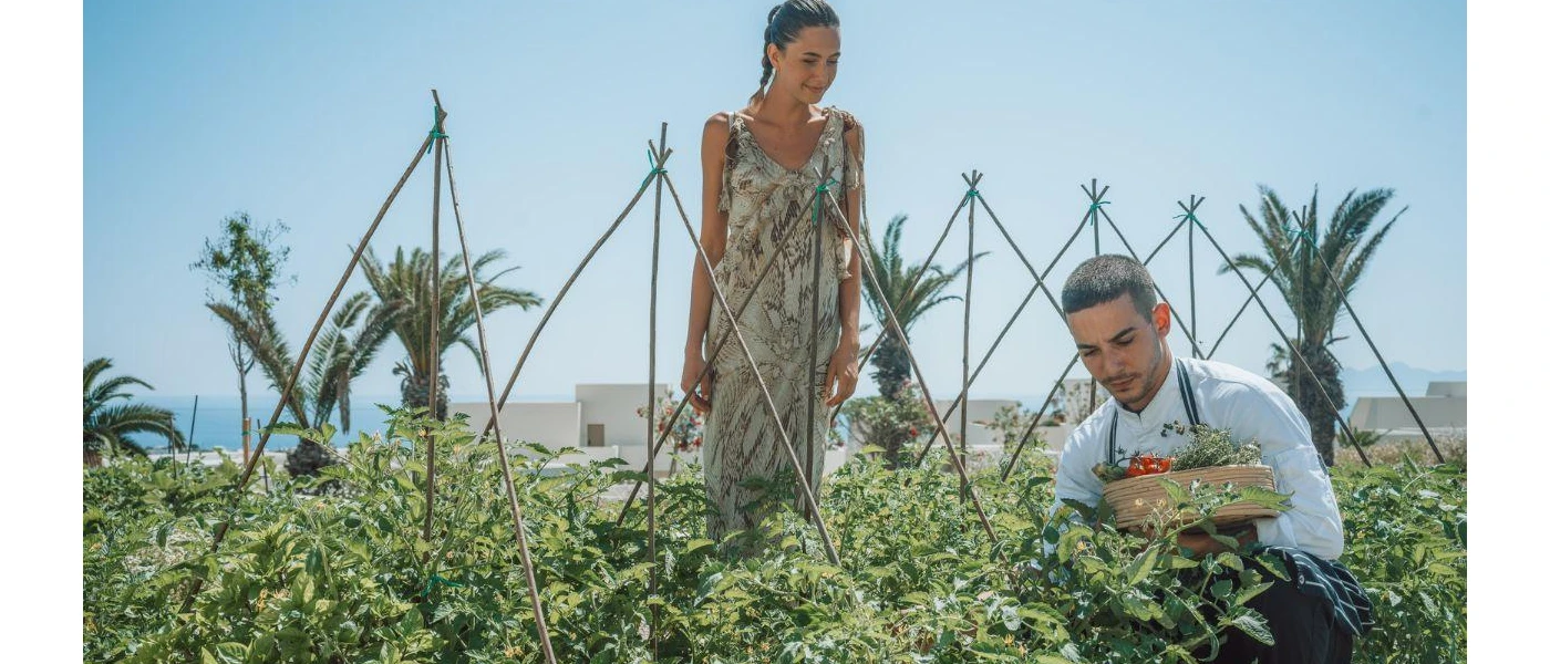 Woman in floral dress watches as a staff member picks herbs and vegetables from a kitchen garden under a blue sky