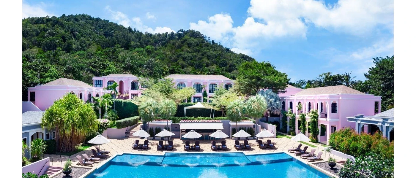 Swimming pool next to a paved terrace with white parasols and palm trees, surrounded by pink buildings