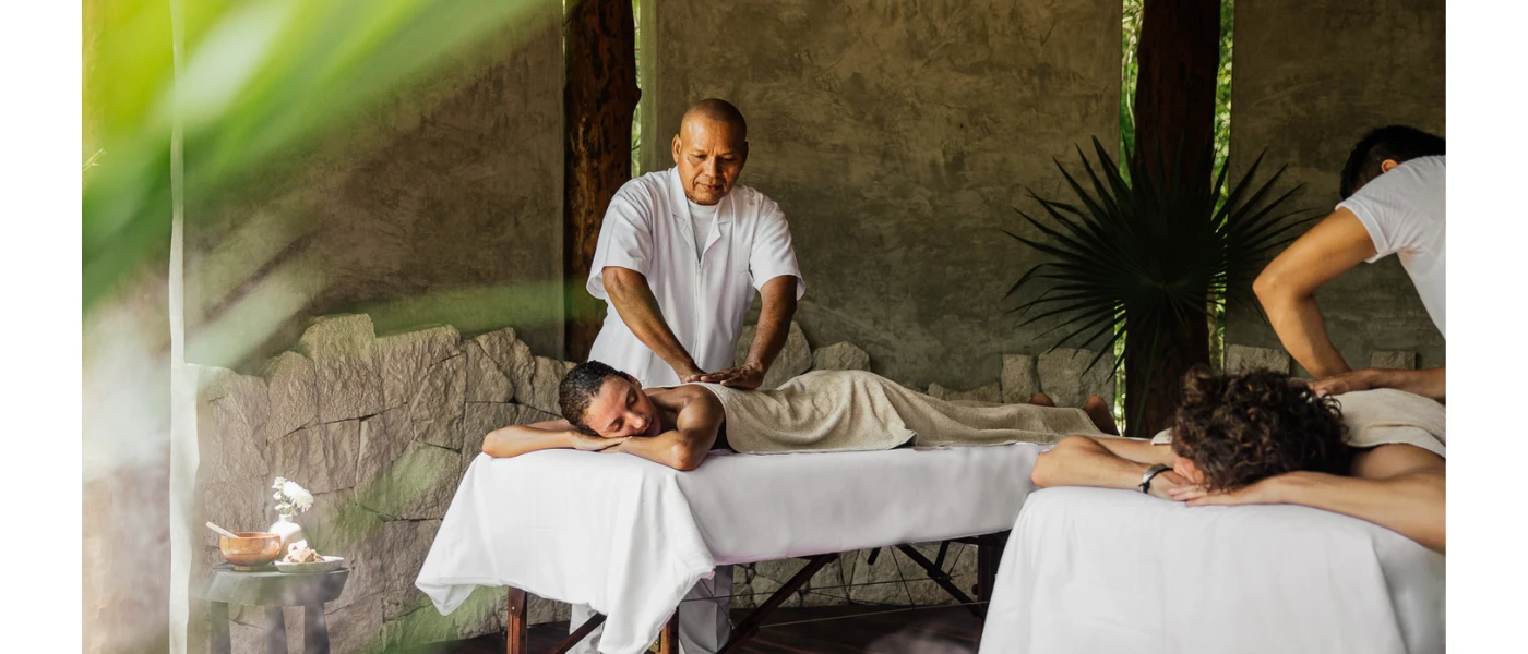 Man and woman lie on their fronts on massage beds in an open-air cabana surrounded by greenery, as two therapists massage their backs