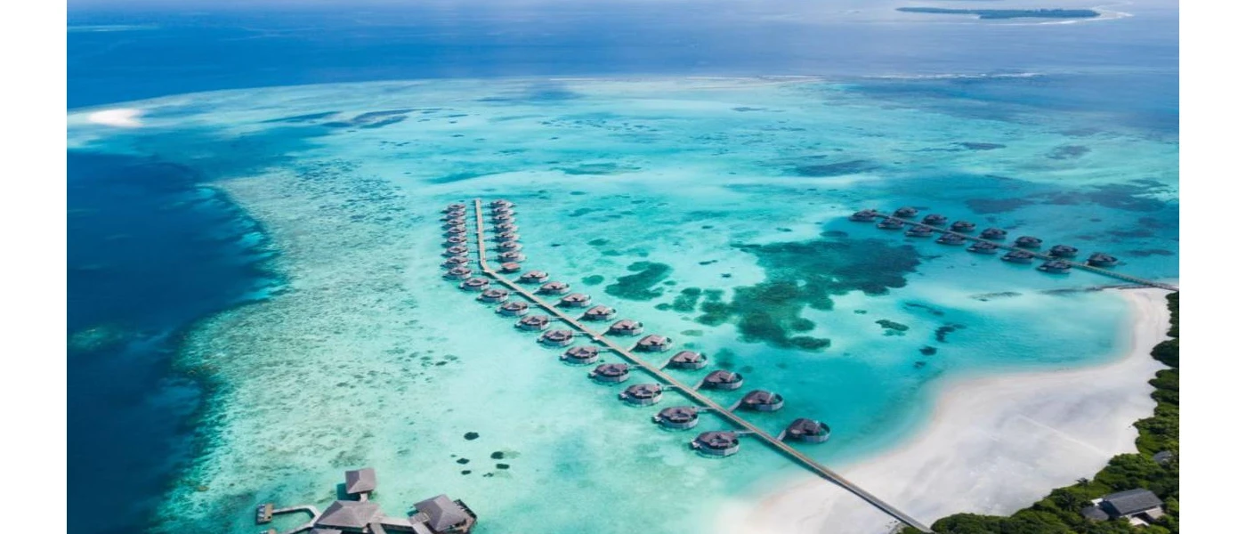 Over-water villas in turquoise shallows in the Maldives, overlooking a white-sand beach and an island with a lush interior