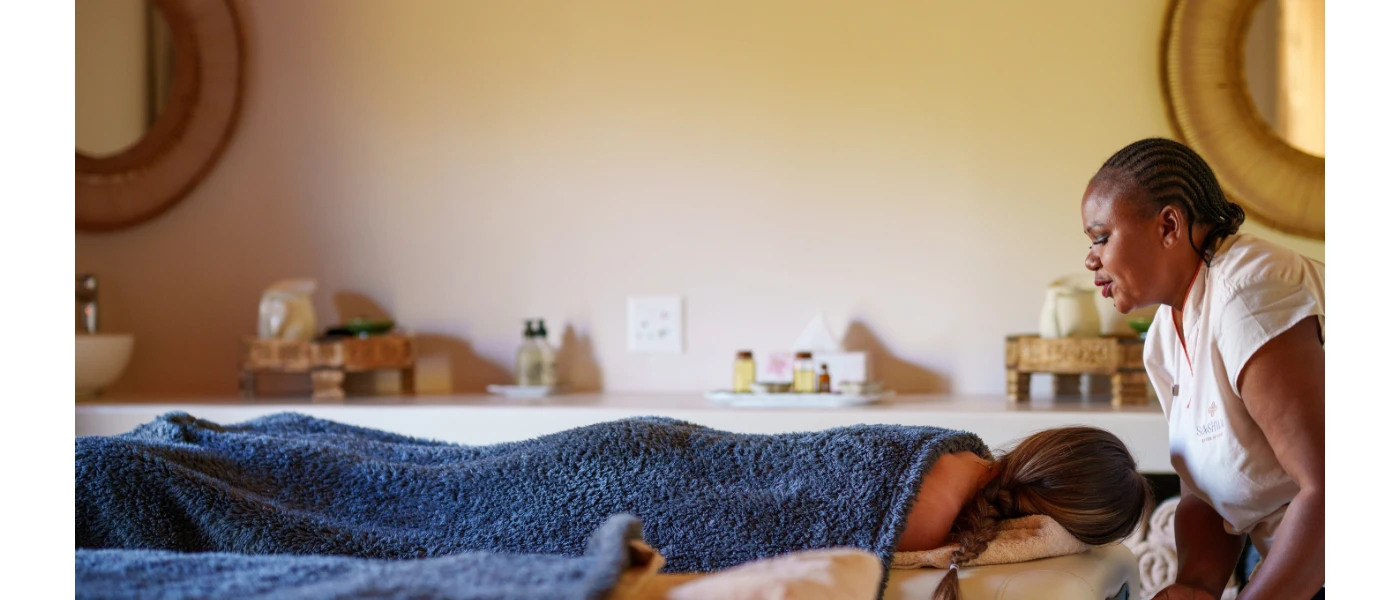 Woman lies on her front under a blue towel as a therapist in white uniform prepares for her spa treatment