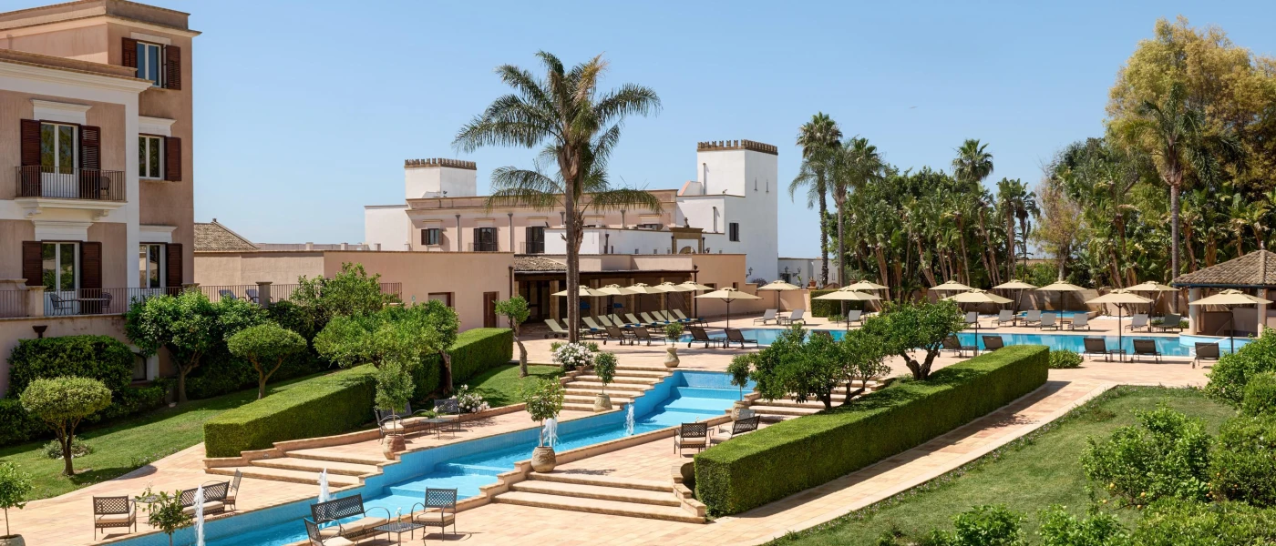 Turquoise fountain designed to look like a pool, which flows over steps in a stone terrace surrounded by gardens