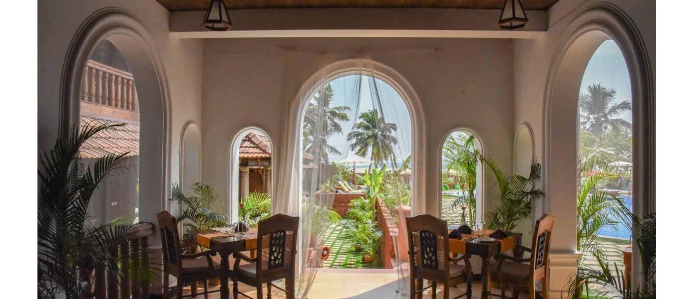 Restaurant with arched doorways, white walls, dark-wood tables and chairs and potted plants overlooking a pool terrace