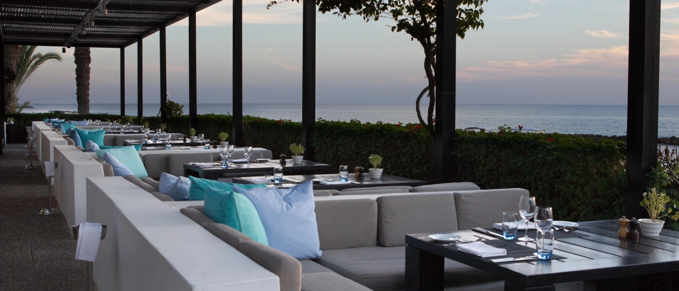 Restaurant with beige cushioned sofas and blue pillows and tables set up for meals awaiting guests, sea view