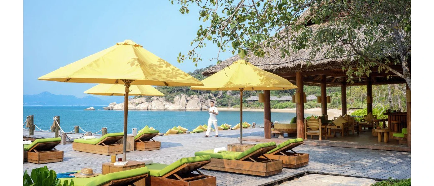 Oceanfront wooden terrace with yellow parasols, green-cushioned loungers and a waiter in white carrying a tray