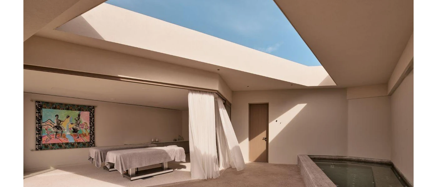 Angular cream-coloured room with an open roof, small pool, massage bed and white curtain, plus a colourful painting on the wall