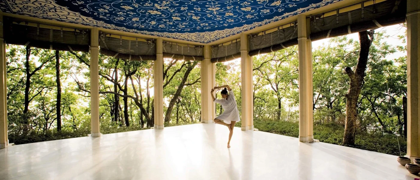 Woman in a white shirt dress in a yoga pose in the middle of an open-air pavilion surrounded by tropical greenery