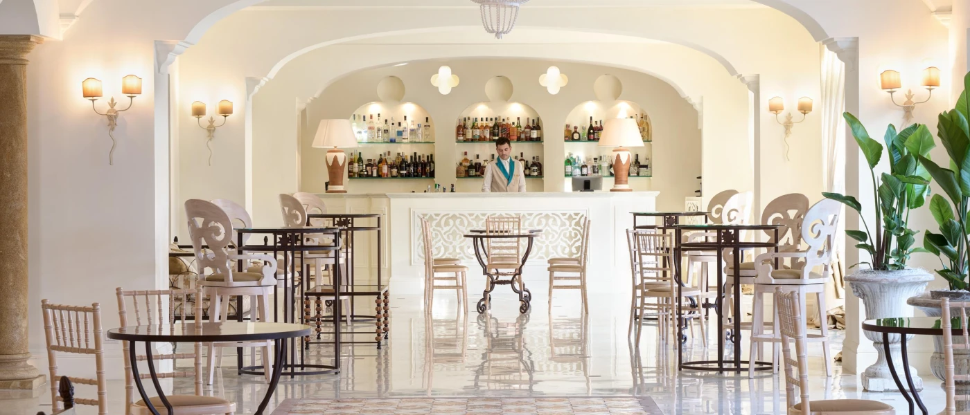 Bright bar with white walls and glossy floors, a man in a cream blazer standing behind the bar, and elegant wall lights