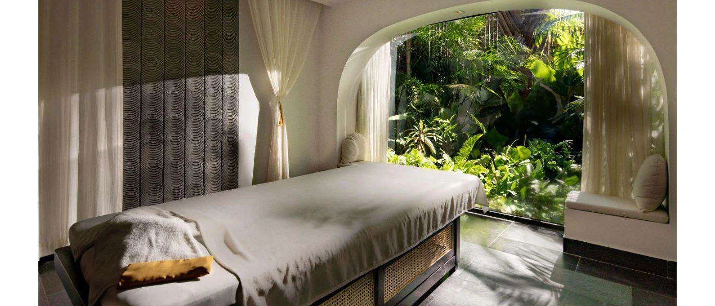 Spa treatment room with a massage bed draped in grey fabric, and a large curved window that looks out to tropical gardens