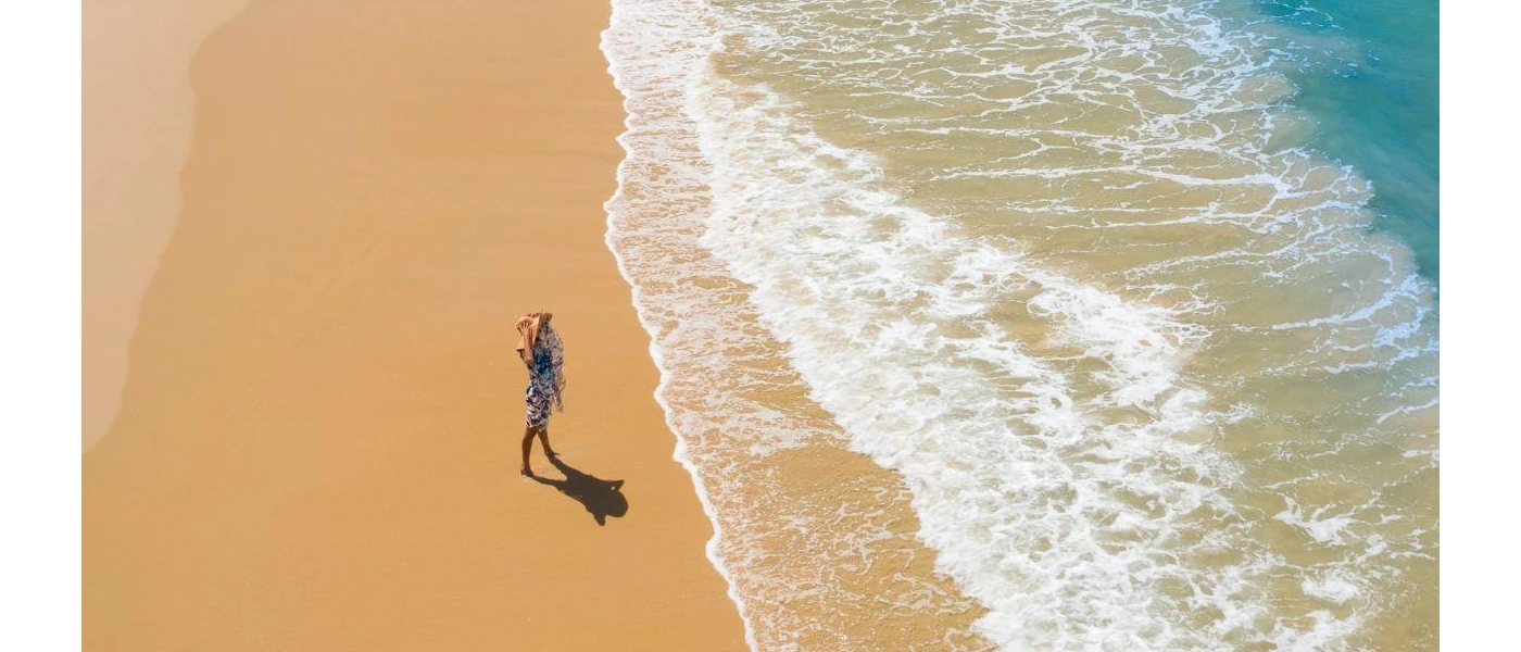 Woman in a straw hat and floral dress stands on the shoreline 
