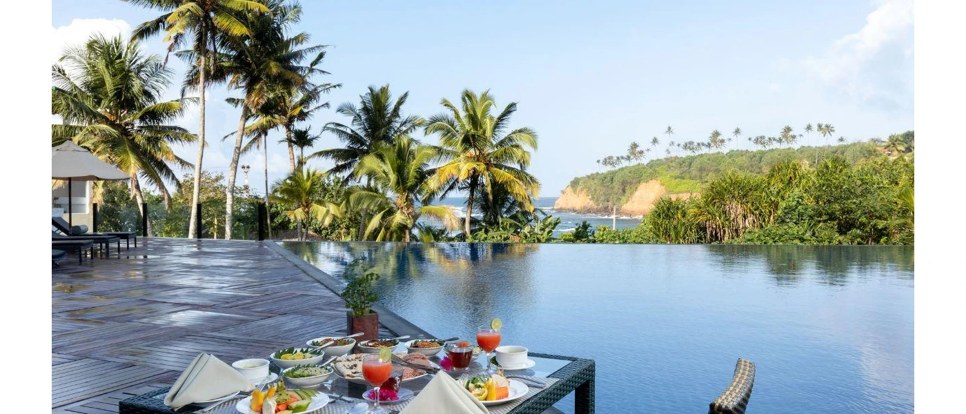 Infinity pool overlooking the sea and tropical gardens with a table next to it laid up with fruit, salads and smoothies