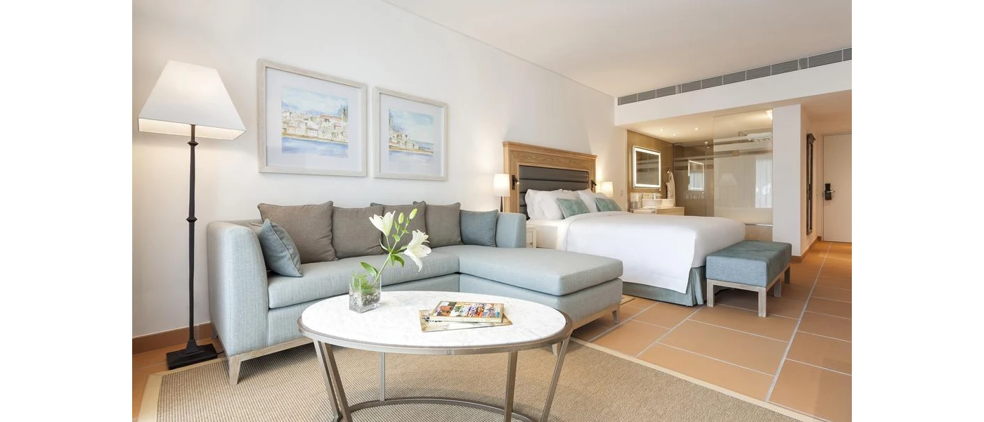 Hotel room with terracotta tiled floors, white walls and grey soft furnishings, including a sofa and cushions, bed pillows and headboard, plus a white coffee table and a floor light with a black stand