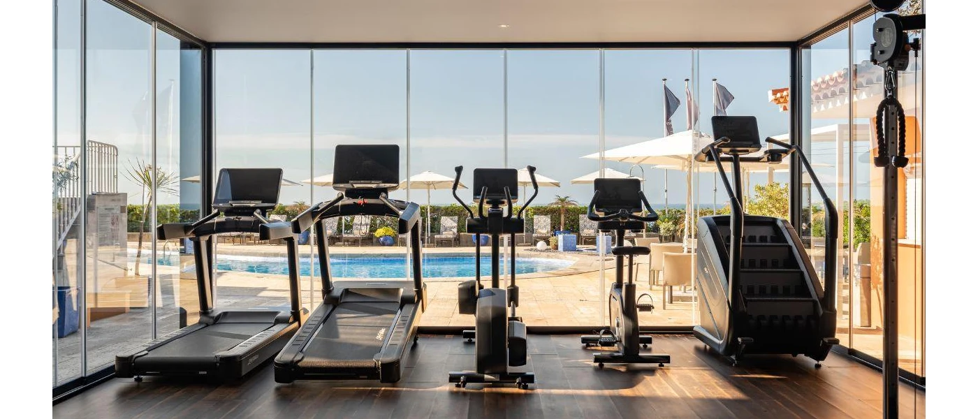 Treadmills, bikes and a stepper in a gym with floor-to-ceiling windows overlooking a pool