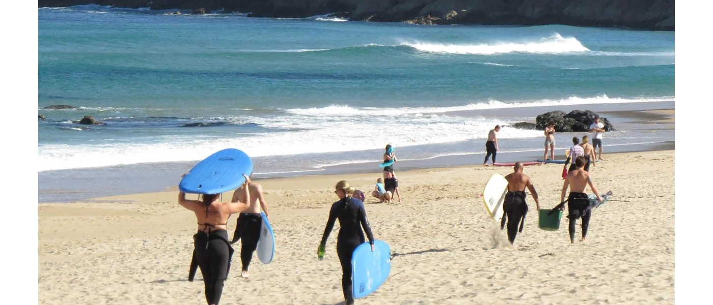 Busy beach will people in wet suits, some carrying surf boards, some body boards and some in the water