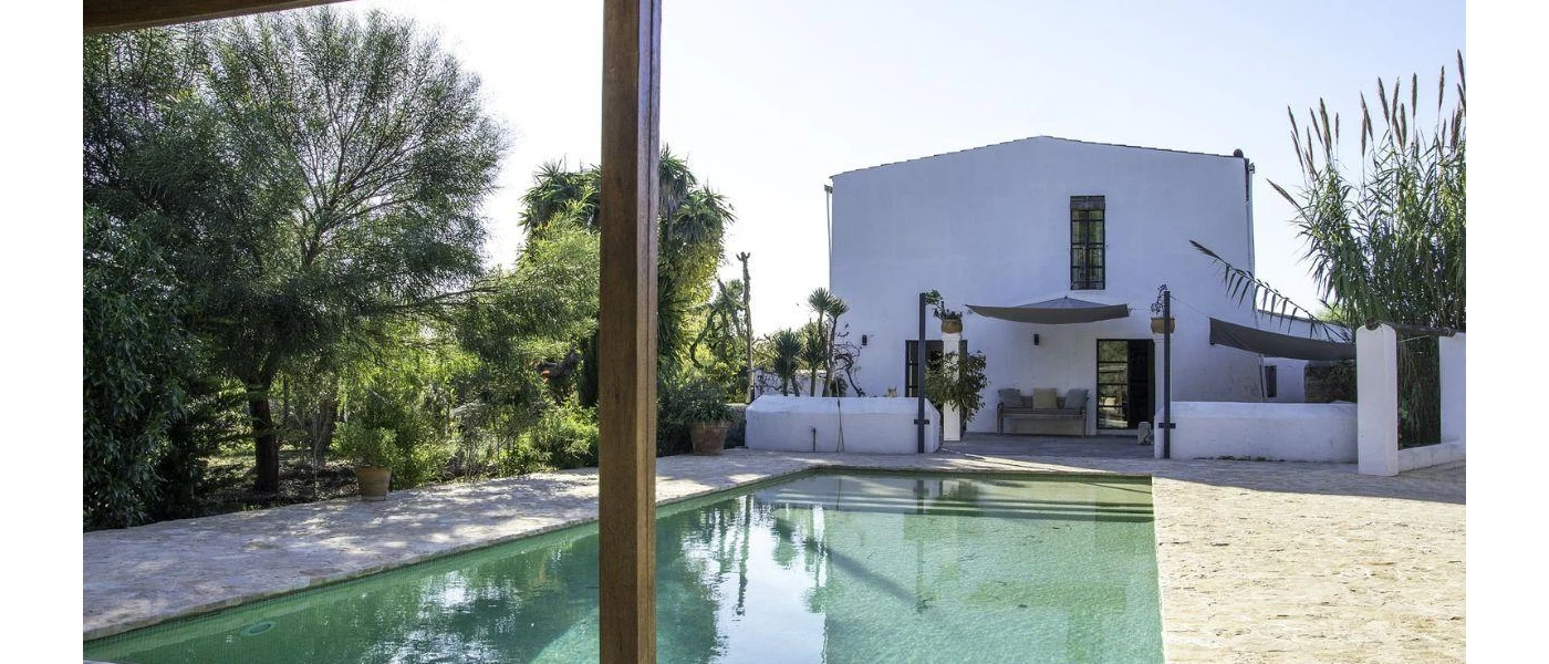 White cube-shaped villa with a rectangular pool, cobbled stone terrace and surrounding greenery