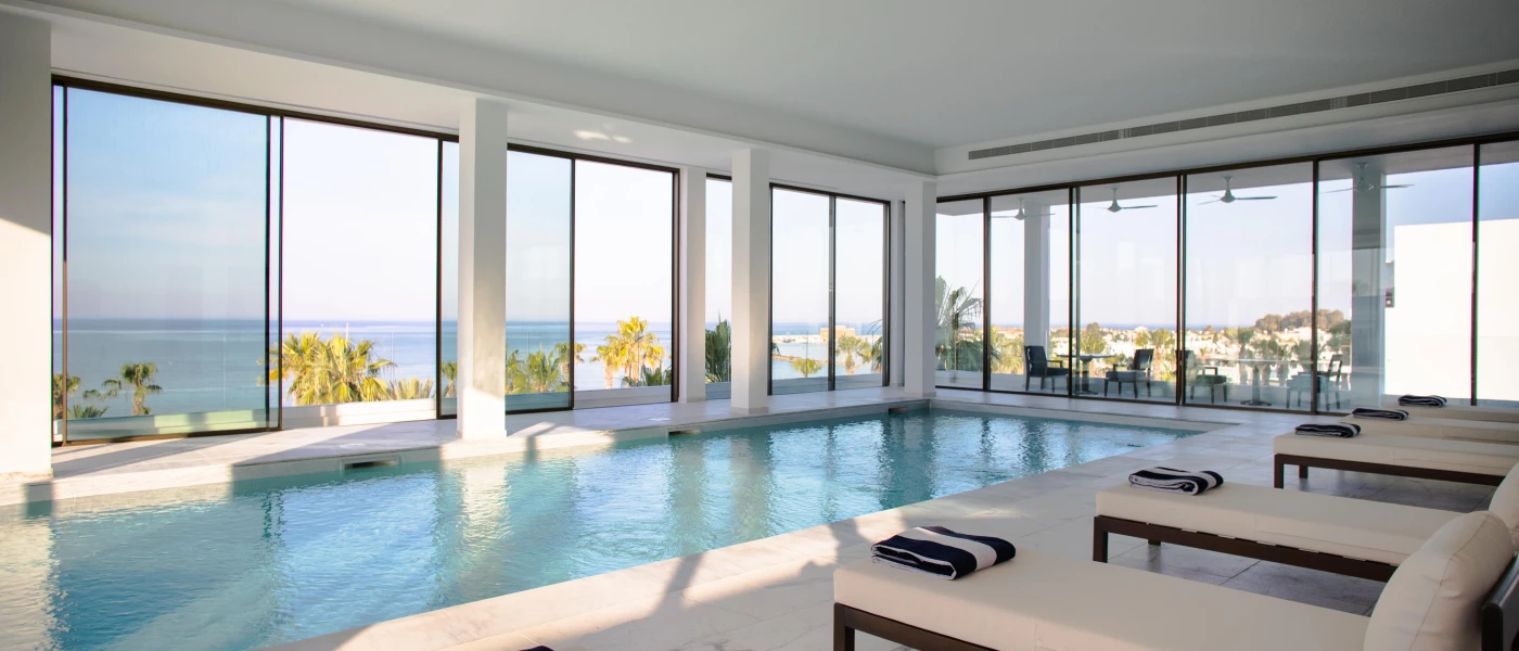 Indoor swimming pool surrounded by white pillars, white-cushioned dark-wood loungers with striped towels on them, and floor-to-ceiling windows with a sea view