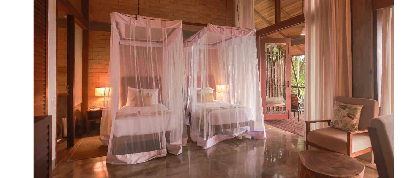 Hotel bedroom with wooden floors and ceiling beams, two beds with white muslin drapes and patio doors opening onto a balcony area