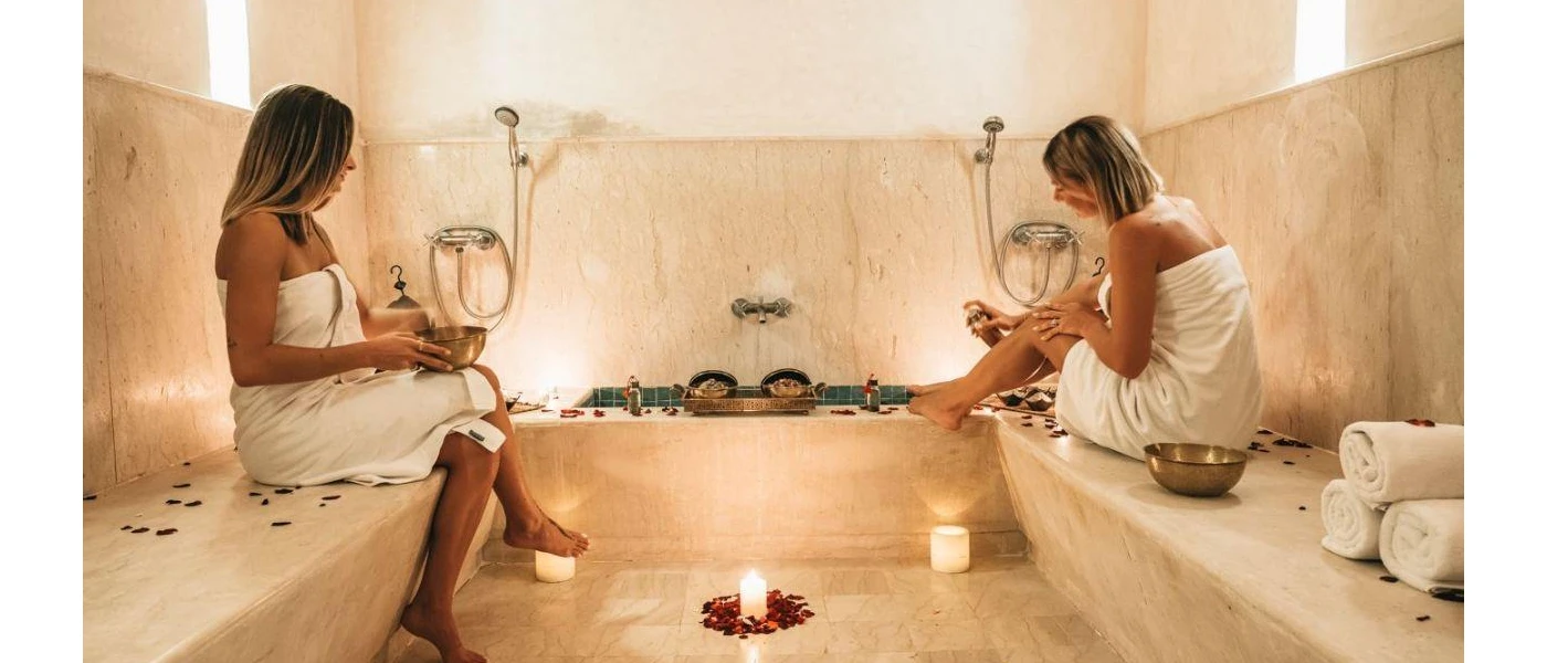 Two women chatting in a steam bath in white towels with candlelight and fluffy white spare towels