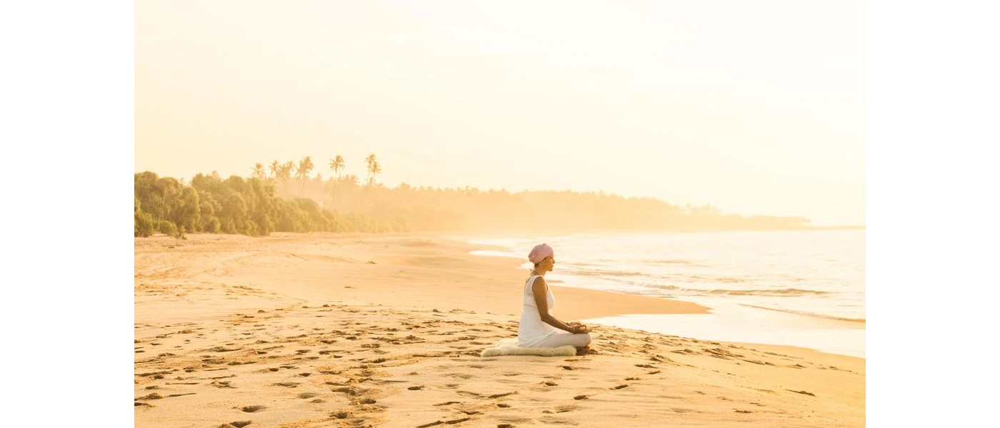 Woman in a white dress and headscarf sitting on a cushion on the beach, gazing out to sea as the sun rises 