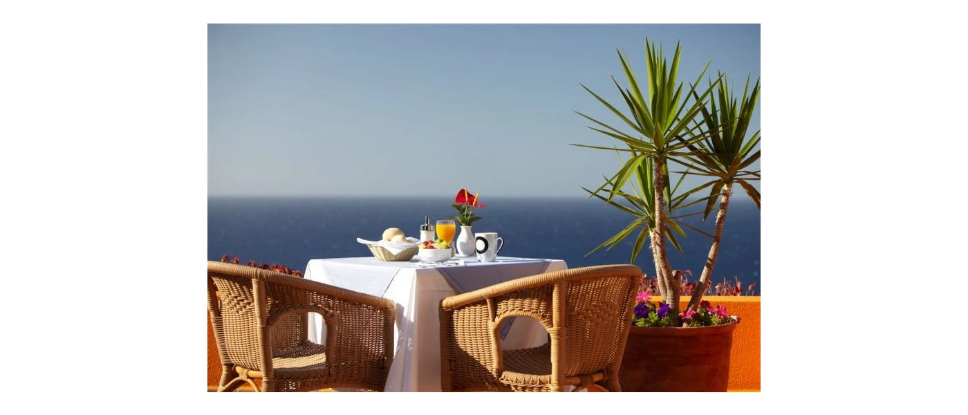 Table with a white tablecloth laid on a sunny terrace overlooking palm trees and the sea
