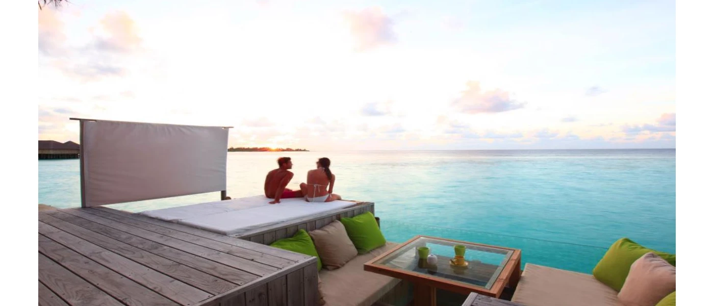 Couple sit on a cushioned terrace area, next to sofas with cream and green cushions overlooking their private pool and the ocean