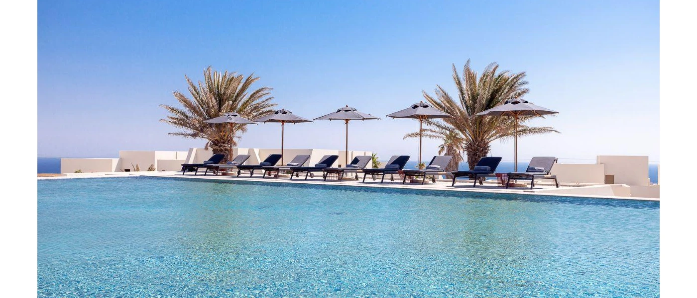 Lake-like swimming pool under a blue sky, edged by palm trees, grey parasols and grey-cushioned loungers