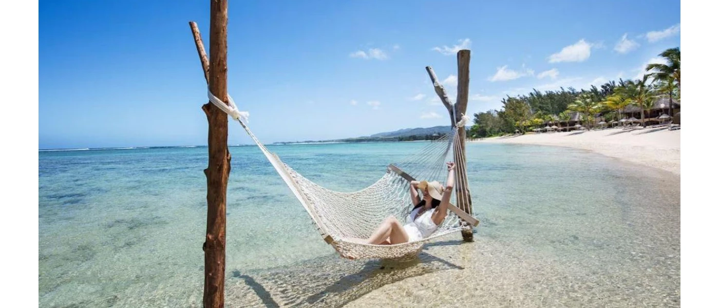 Woman in a white swimsuit and a straw hat lying in a hammock in clear shallows next to a sandy beach