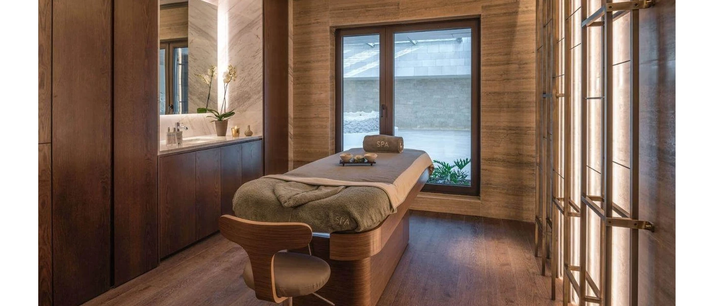 Wood-panelled spa treatment room with a massage bed and neighbouring sink and mirror, plus a large window behind