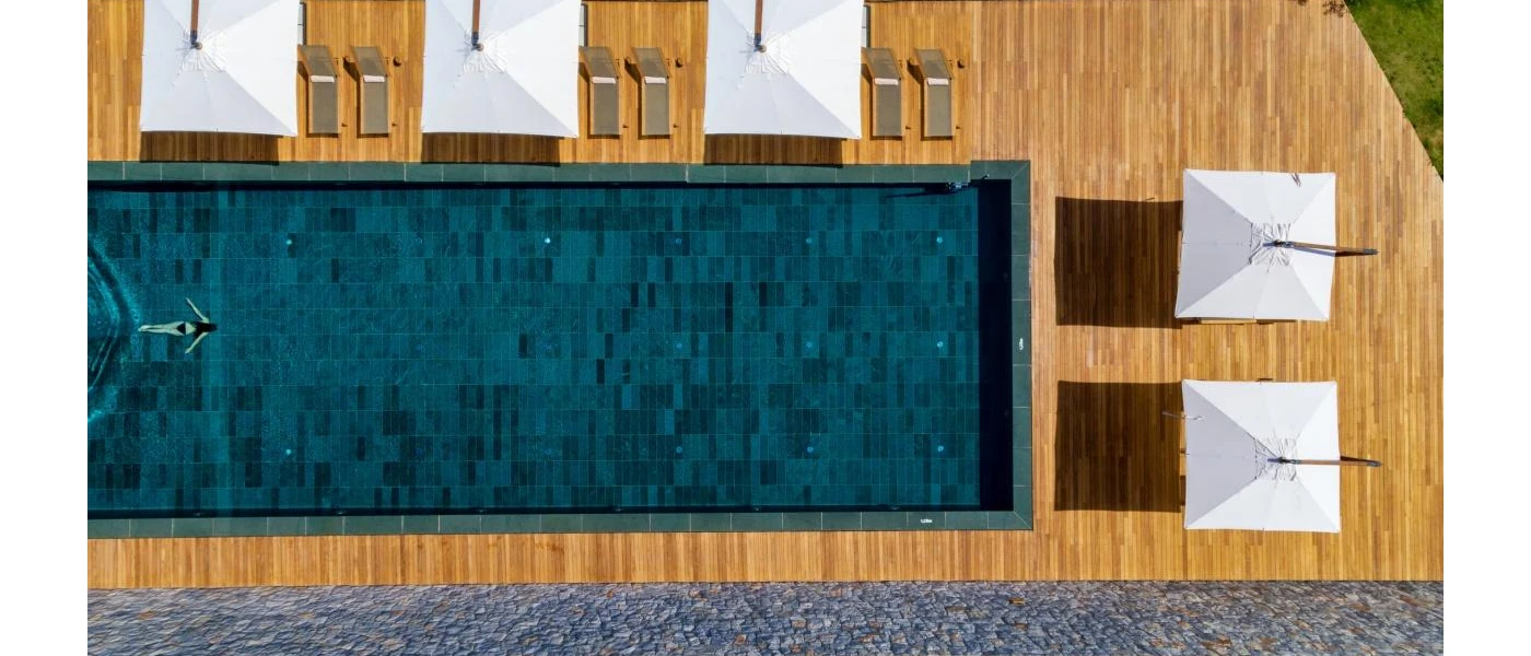 Aerial view of a women in a black swimsuit swimming in a rectangular pool, surrounded by sleek wooden decking and white parasols