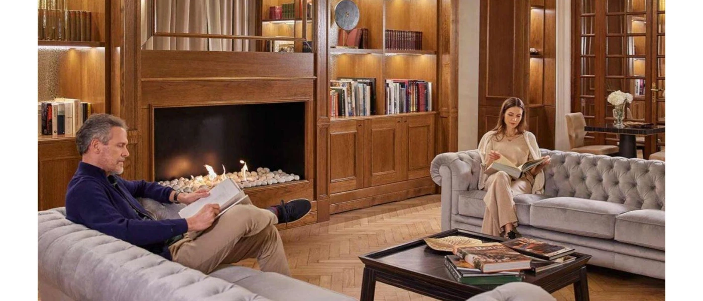 Wood-panelled room with a fireplace, bookshelves, and two people reading on grey chesterfield-style sofas