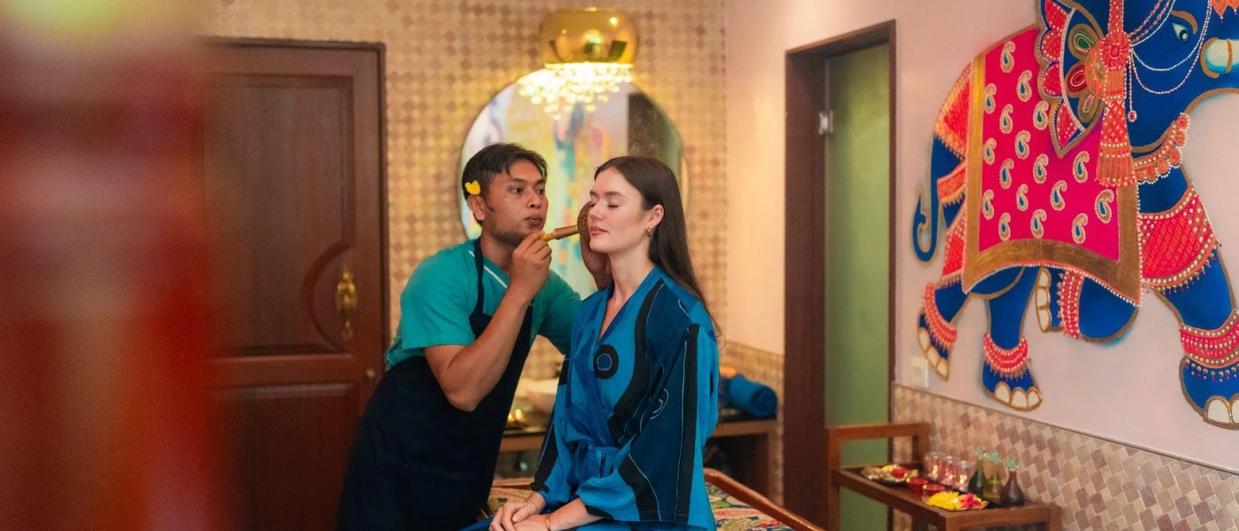 Therapist in an apron examining a robed woman in a treatment room with an elephant mural on the wall