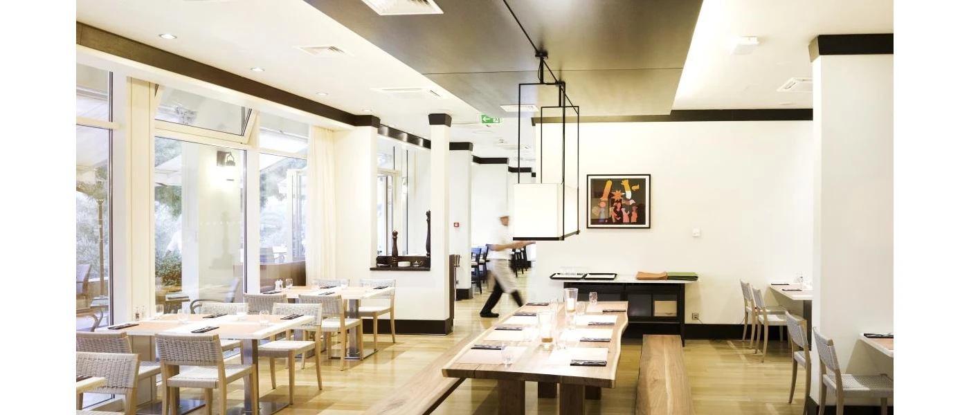 Minimalist restaurant with white walls, wooden tables and bright lighting, as a chef carries something in the background