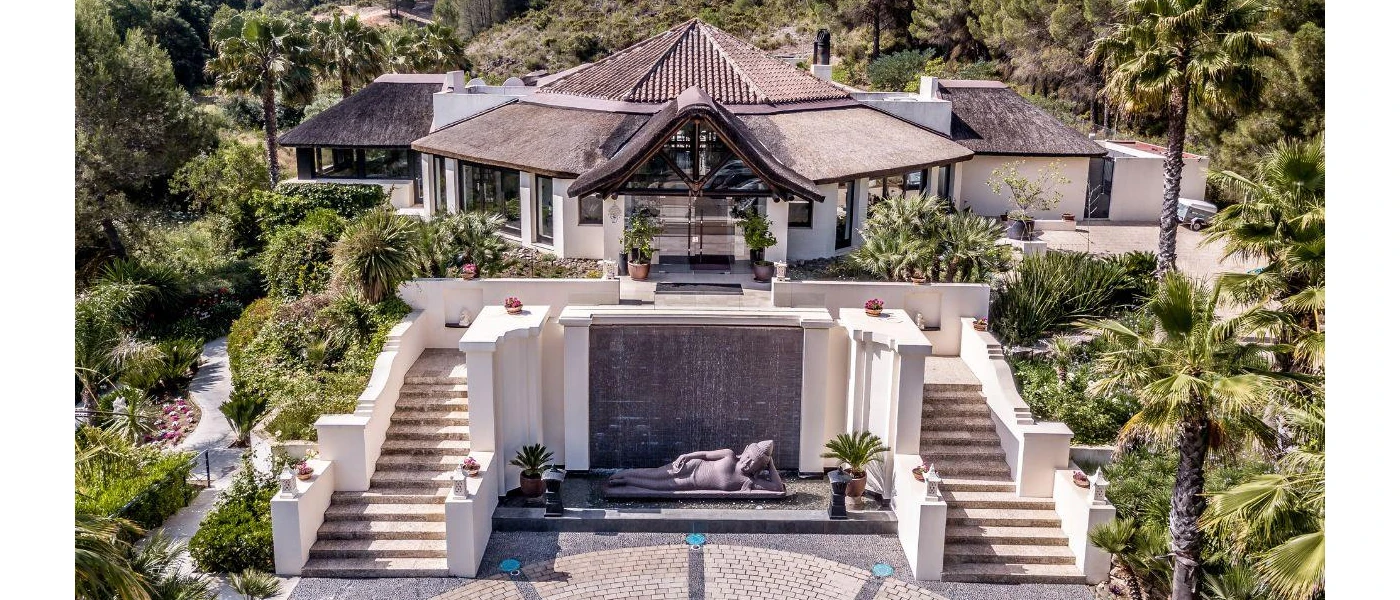 Stylish resort facade, with a low-rise building with a triangular rooftop, symmetrical steps leading down to a lower level with a waterfall and a Buddha statue, and surrounding tropical gardens