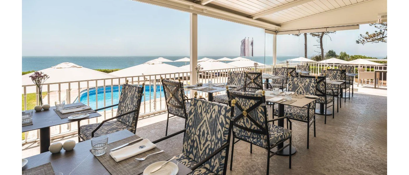 Outdoor restaurant terrace with blue and white patterned chairs and a cream cover with a view of the pool and ocean